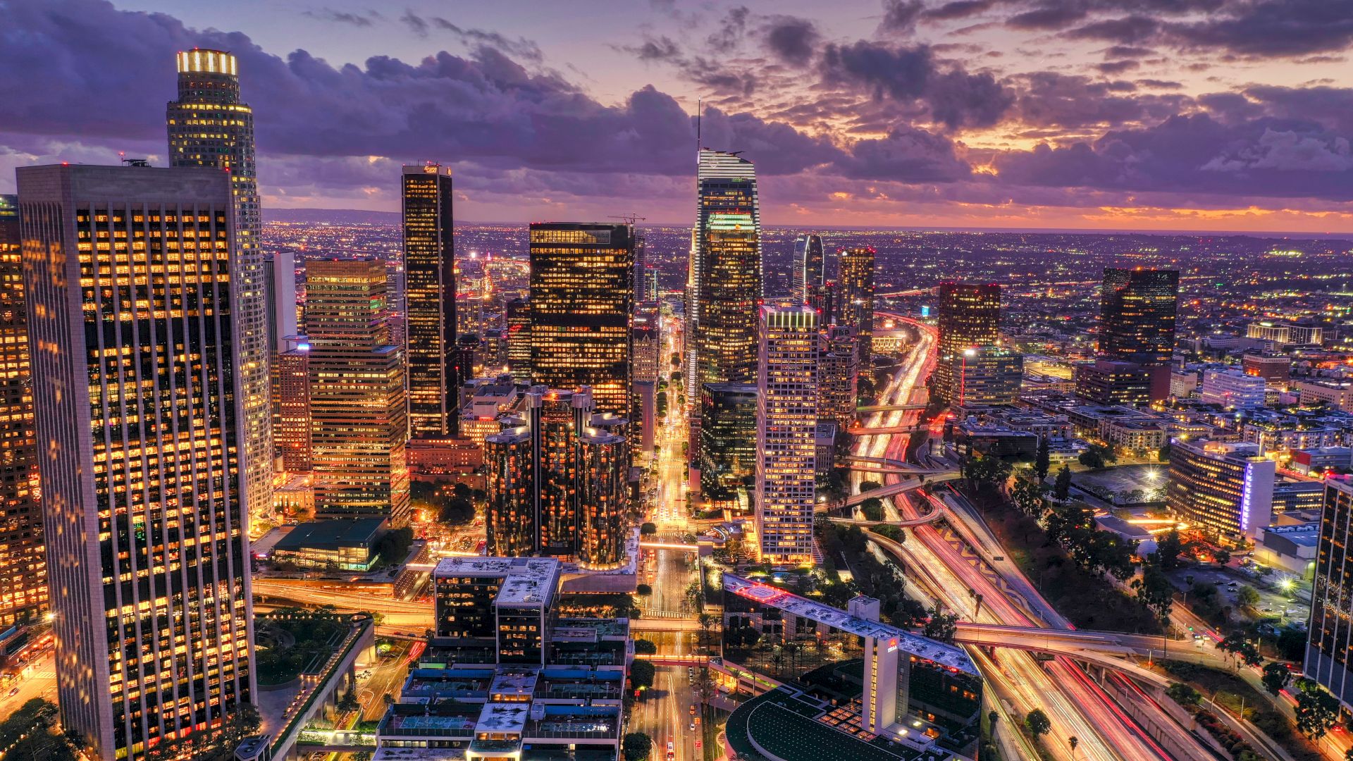 A vibrant city skyline at dusk with illuminated skyscrapers and busy streets, framed by colorful clouds and a fading orange-purple sky.