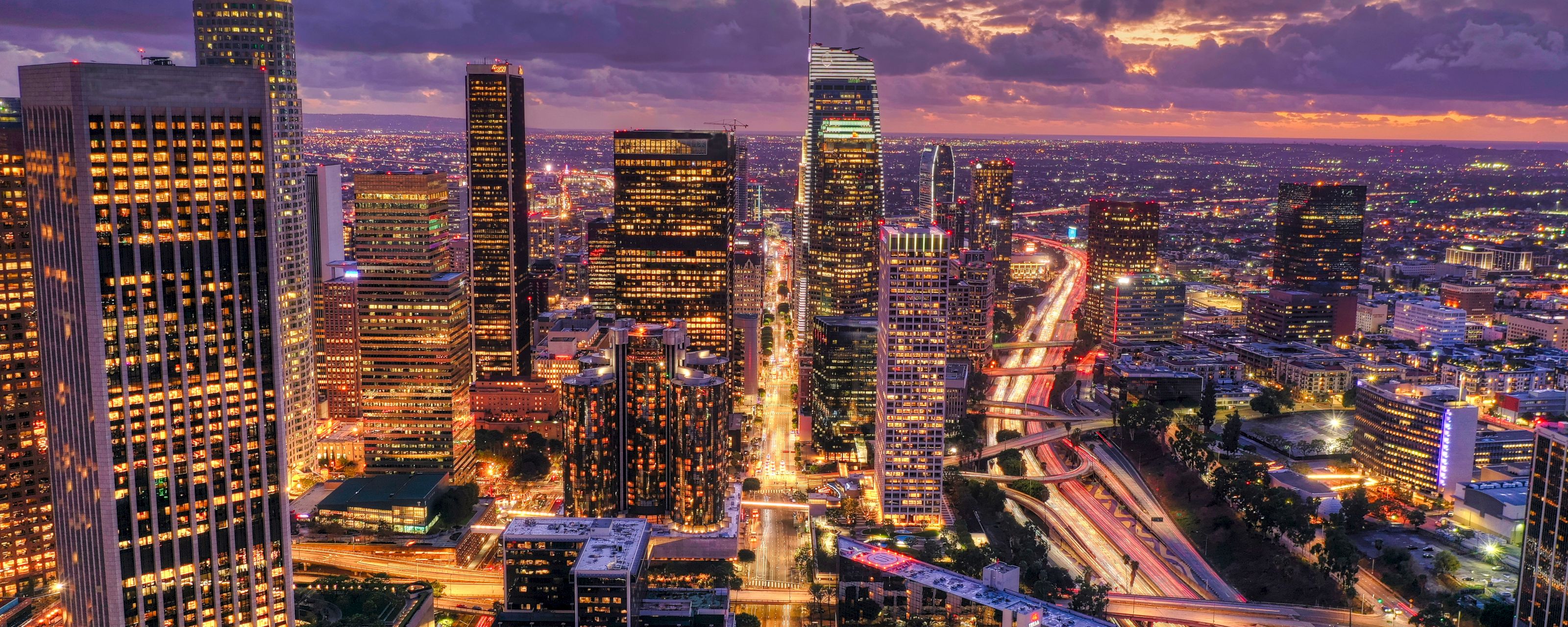 A cityscape at sunset with skyscrapers and a highway.