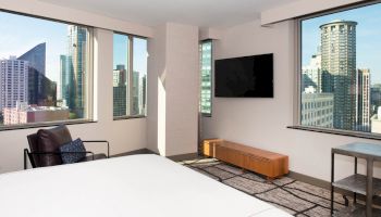 The image shows a modern hotel room with large windows and a view of city skyscrapers, featuring a bed, chair, TV, and minimalistic decor.