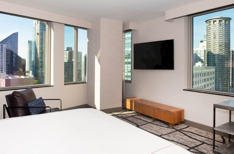 The image shows a modern hotel room with large windows and a view of city skyscrapers, featuring a bed, chair, TV, and minimalistic decor.