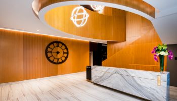 A modern lobby with a marble reception desk, wooden wall, large clock, spiral staircase, hanging lights, and a vase of flowers ending the sentence.
