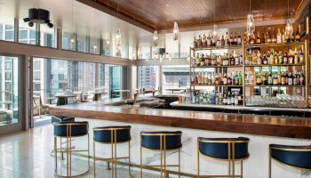 Modern bar with high ceilings, stylish lighting, and large windows, showcasing a well-stocked bar and seating area with five blue bar stools.