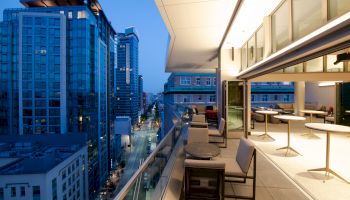 A modern cityscape with high-rise buildings at dusk, viewed from a balcony with tables and chairs, overlooking a street lined with more buildings.