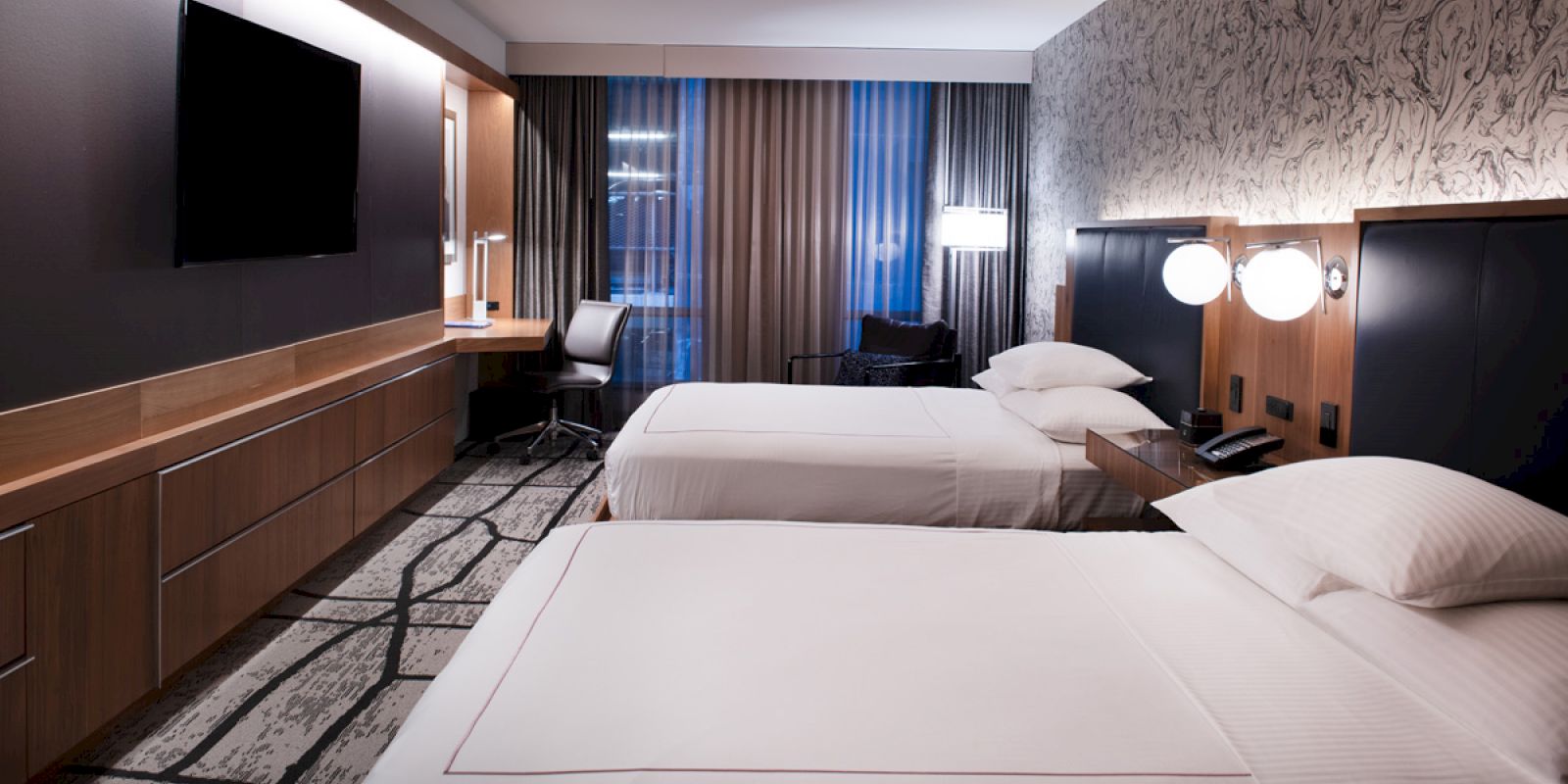 A modern hotel room with two beds, a flat-screen TV, a desk with a chair, and contemporary decor including patterned wallpaper and stylish lighting.