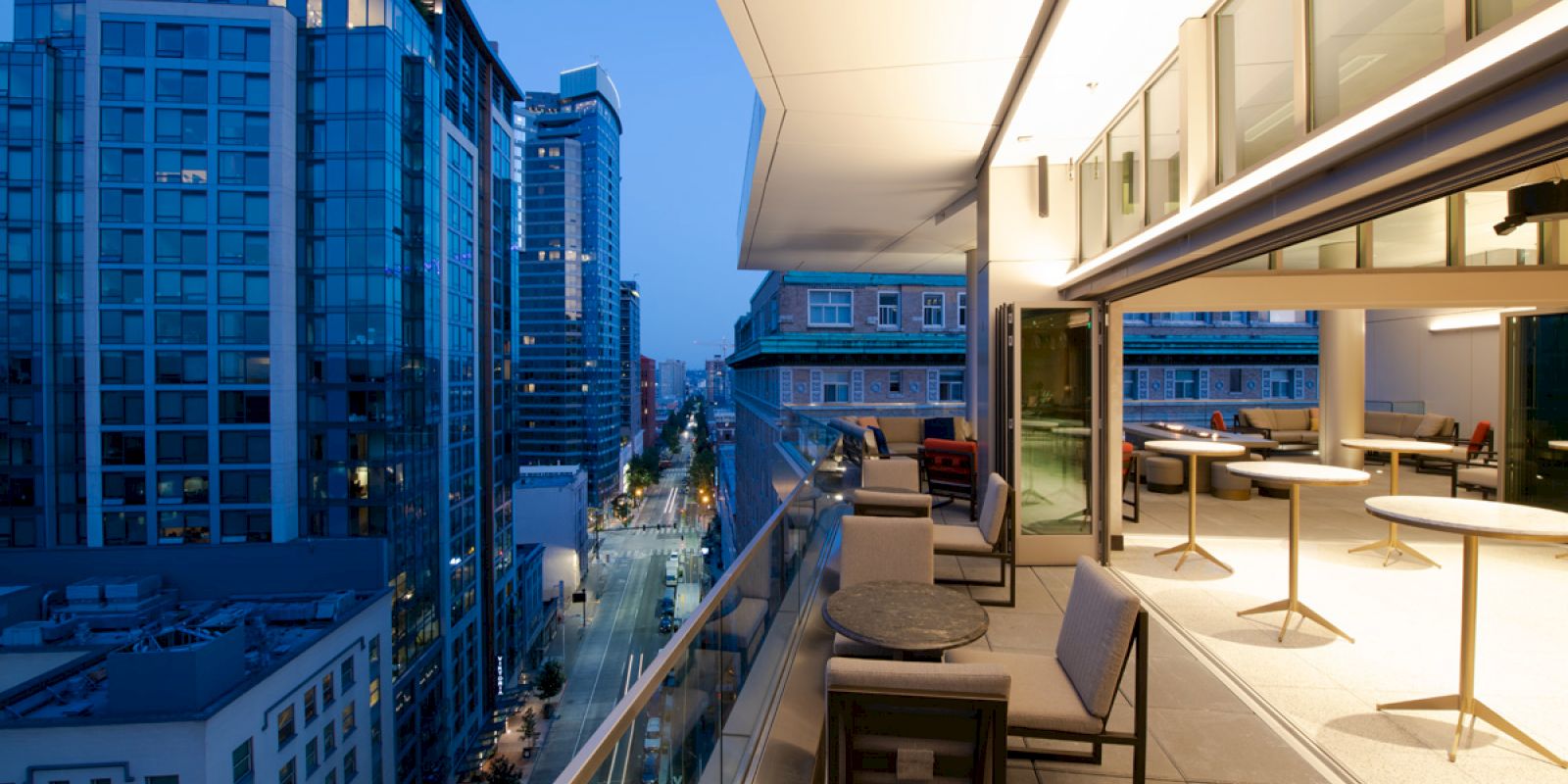 A modern cityscape is seen from a high-rise balcony with tables, chairs, and outdoor seating, overlooking a street lined with tall buildings at dusk.