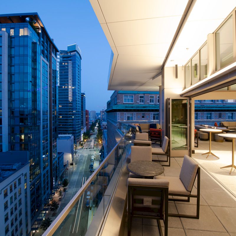 A rooftop terrace with seating overlooks a city street filled with tall, modern buildings, captured during twilight hours.