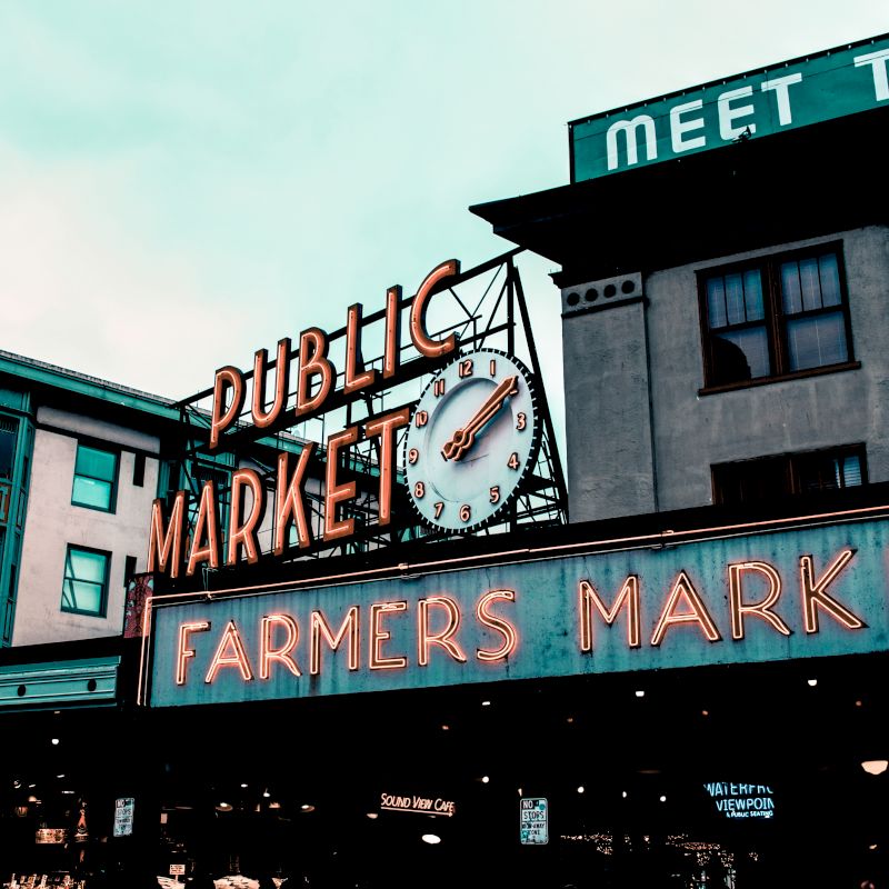 This image shows the iconic neon signs of a "PUBLIC MARKET" and "FARMERS MARKET" with a clock on a building, likely a well-known market location.
