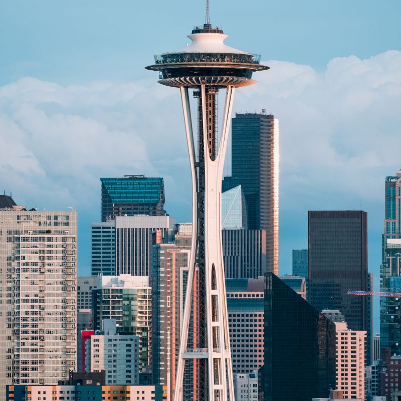 This image shows the Seattle Space Needle with a backdrop of modern skyscrapers and buildings, all under a lightly cloudy blue sky.