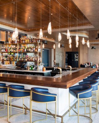 The image depicts a modern bar with a U-shaped counter, blue and gold barstools, overhead pendant lights, and a well-stocked bar area in the background.