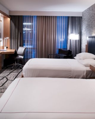 The image shows a modern hotel room with two beds, a wall-mounted TV, a desk with a chair, and a large window with curtains.