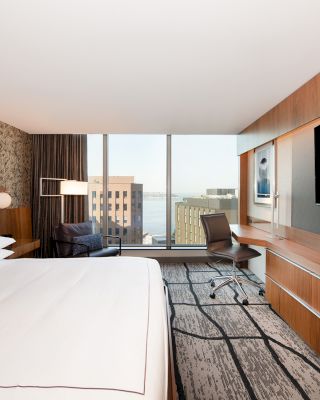 A modern hotel room features a large bed, a wall-mounted TV, a desk with a chair, and a window with a city view and water in the distance.