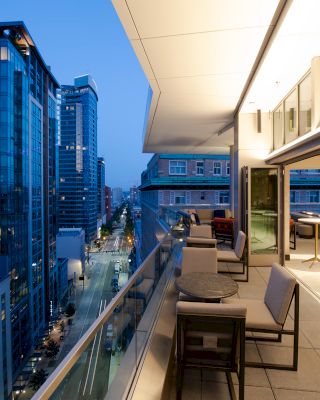 A modern balcony with seating overlooks a city street lined with tall buildings, captured at dusk with the interior space illuminated.