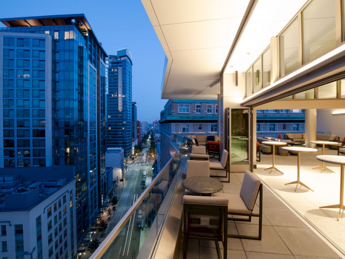 The image shows a modern urban balcony at dusk with seating and tables, overlooking city buildings and a street with lights on.