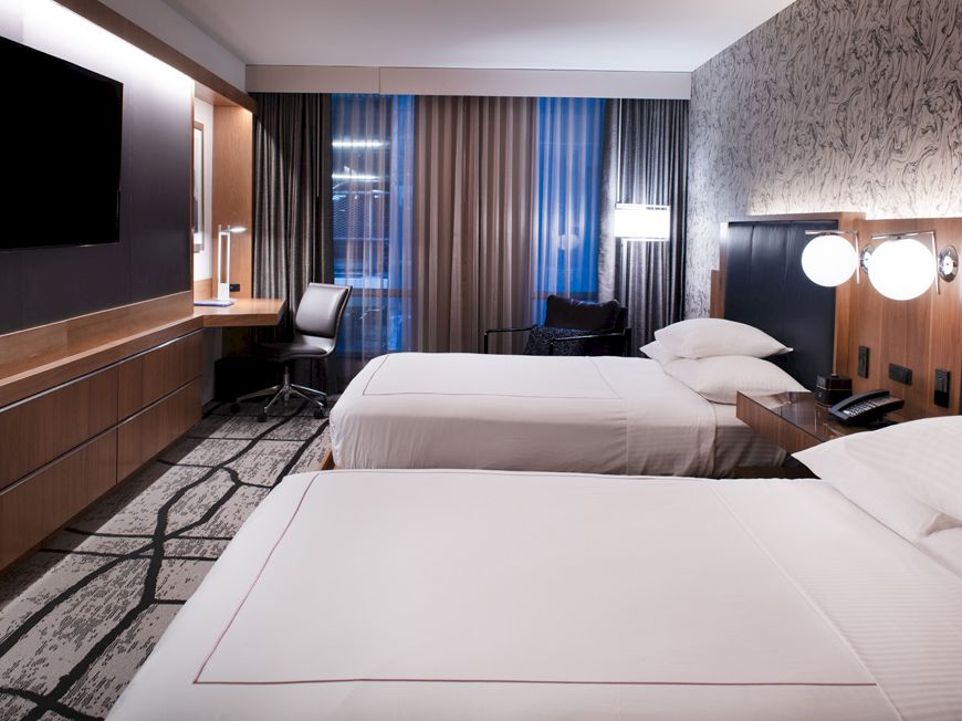 A modern hotel room with two beds, a wall-mounted TV, a desk and chair by the window, and stylish decor and lighting.