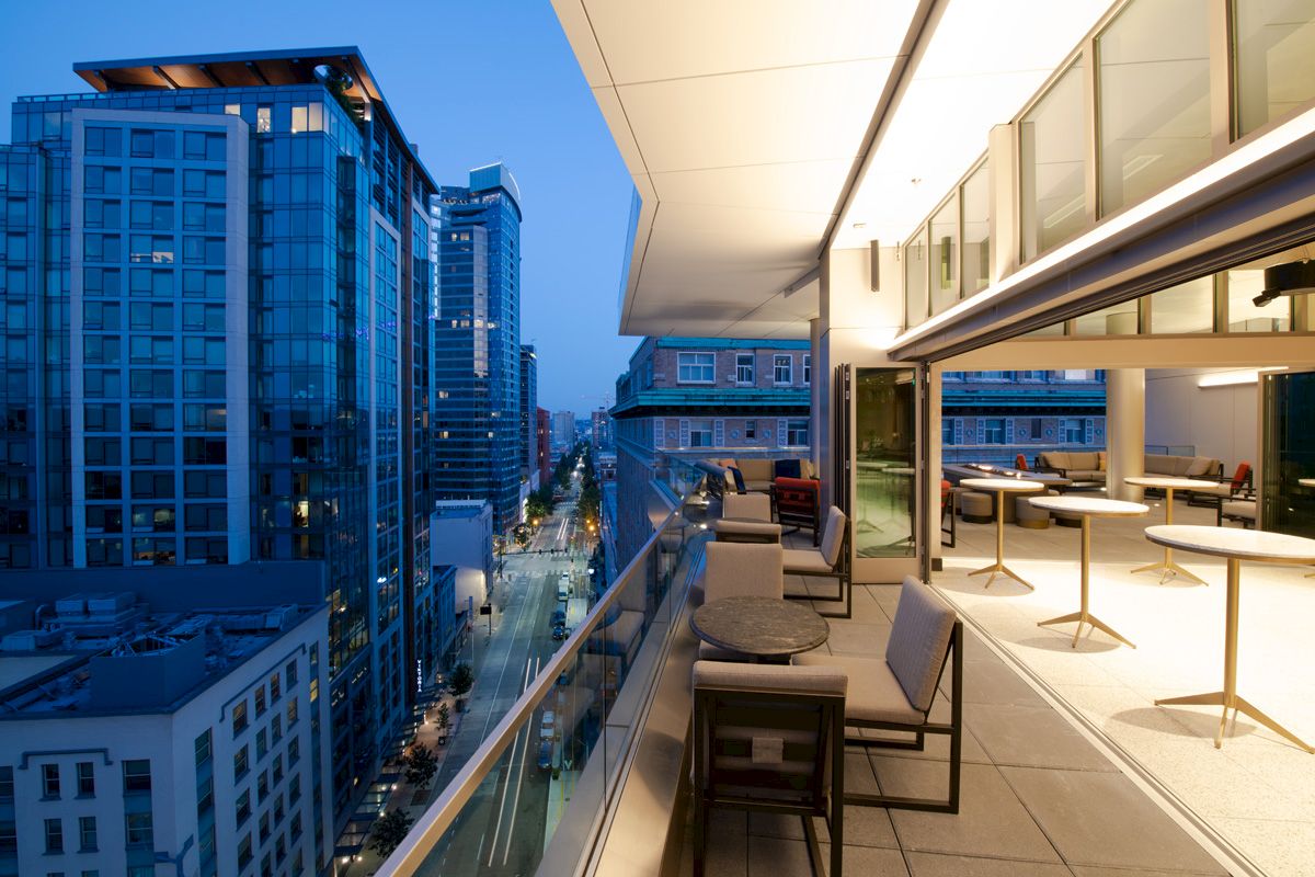 A modern balcony with outdoor seating overlooks a cityscape of high-rise buildings and a street illuminated at dusk.