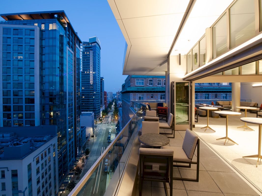 A modern balcony with outdoor seating overlooks a cityscape of high-rise buildings and a street illuminated at dusk.