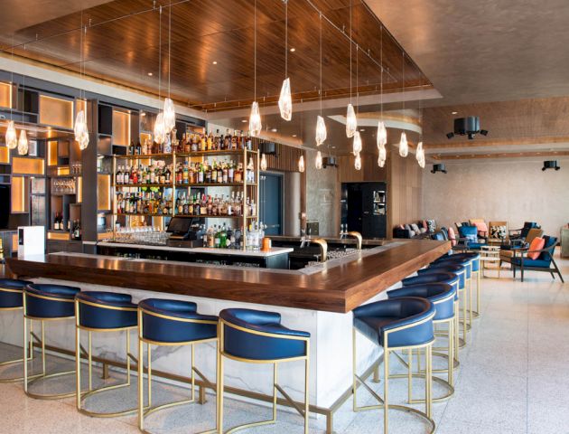 This image shows a modern bar with blue stools, lots of hanging lights, a well-stocked back bar, and a stylish lounge area in the background.