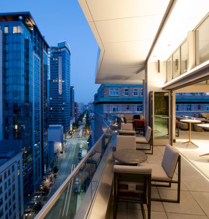 An outdoor balcony with tables and chairs, offering a view of modern high-rise buildings and a city street below, at dusk.