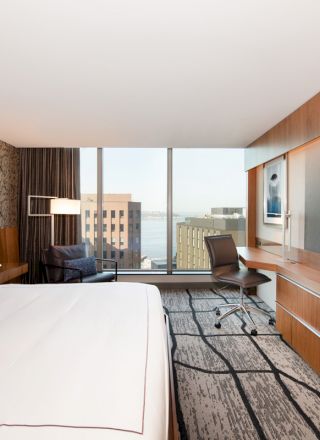 A modern hotel room with a king-sized bed, large window with city view, desk with chair, wall-mounted TV, and contemporary furnishings.