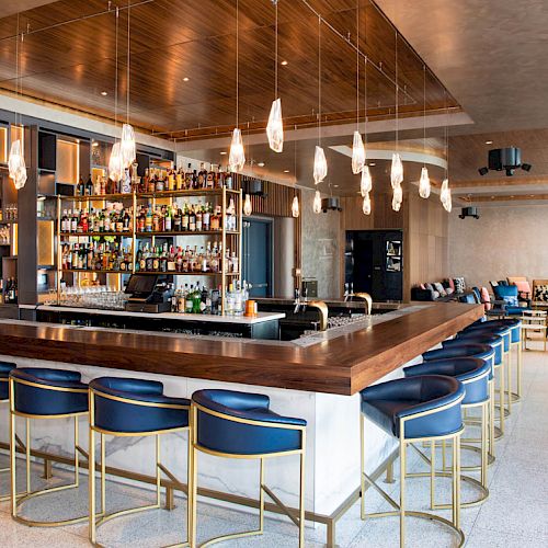 The image displays a modern bar with blue stools, pendant lights, and a variety of bottles behind the counter in a stylish, spacious setting.