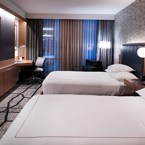 A modern hotel room with two neatly made double beds, a large flat-screen TV on the wall, a desk and chair, and stylish lighting.