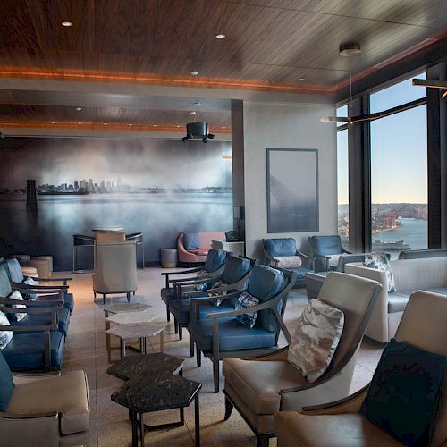 A modern lounge with comfortable seating, large windows offering a scenic view of a coastal city skyline and a ferris wheel.