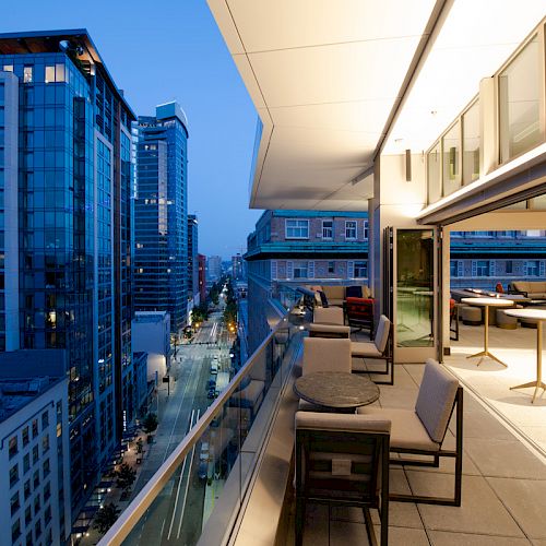 The image shows a modern cityscape at dusk with tall buildings and a balcony featuring seating and tables overlooking the city street below.