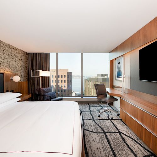 A modern hotel room with a large bed, a wall-mounted TV, a desk and chair, and floor-to-ceiling windows offering a cityscape and partial water view.