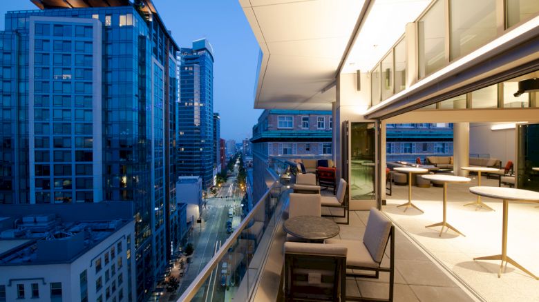 The image shows a modern balcony with seating and tables, overlooking a city with tall buildings. The scene is taken at dusk, with city lights visible.