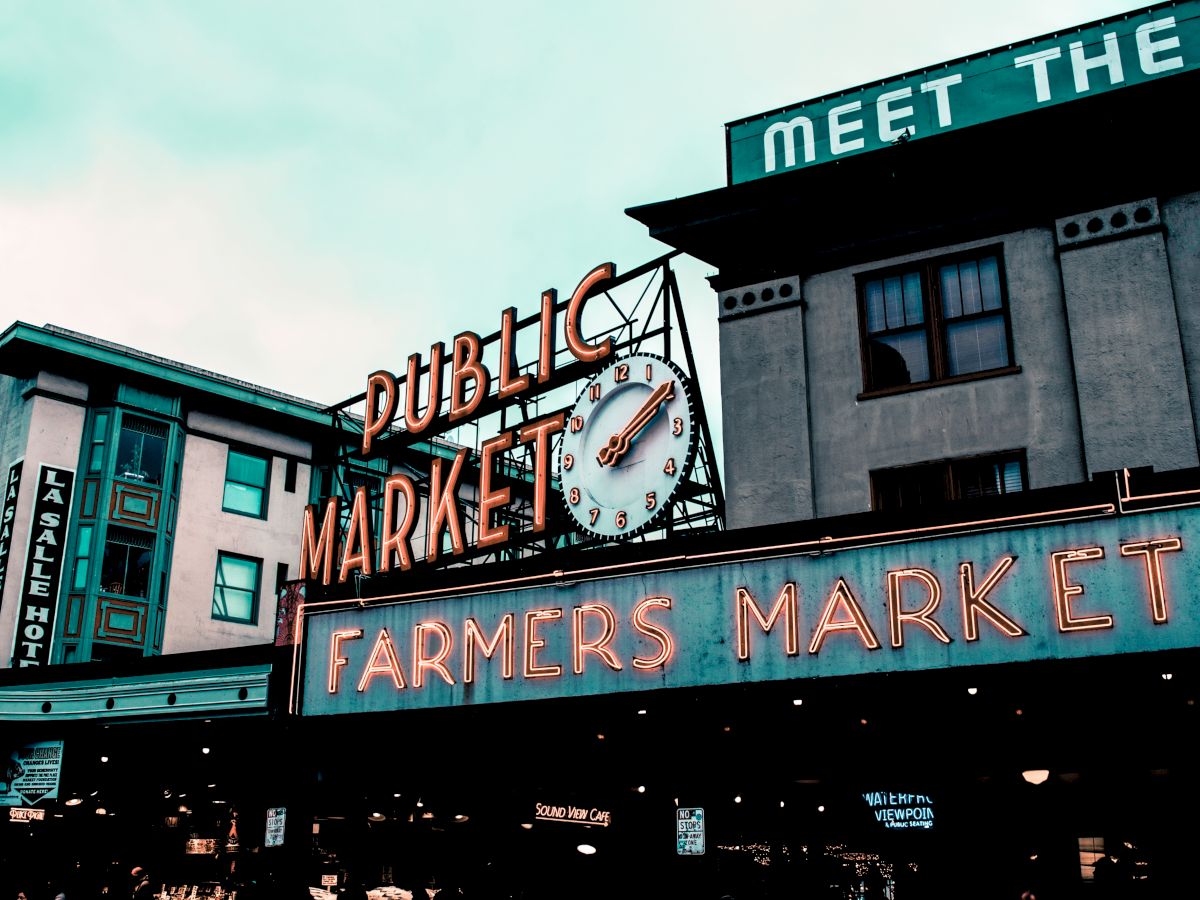 The image depicts the entrance to a Public Market with a clock and a neon sign that reads "FARMERS MARKET," with buildings in the background.