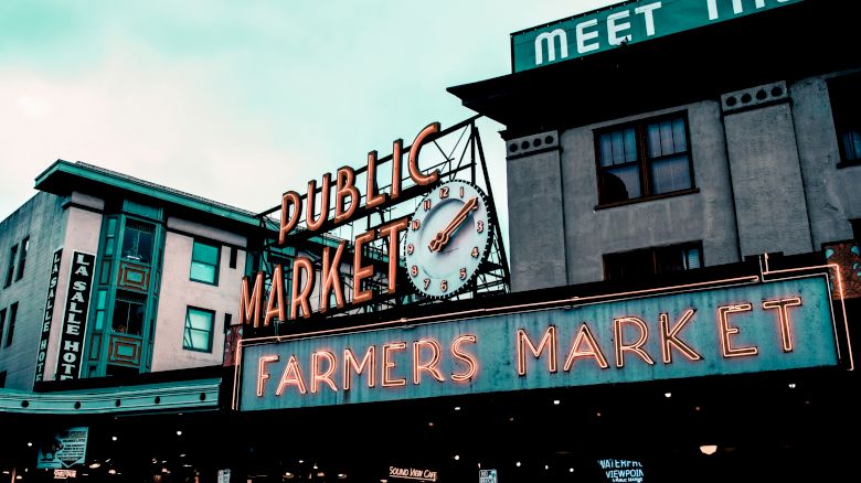 The image depicts the entrance to a Public Market with a clock and a neon sign that reads "FARMERS MARKET," with buildings in the background.