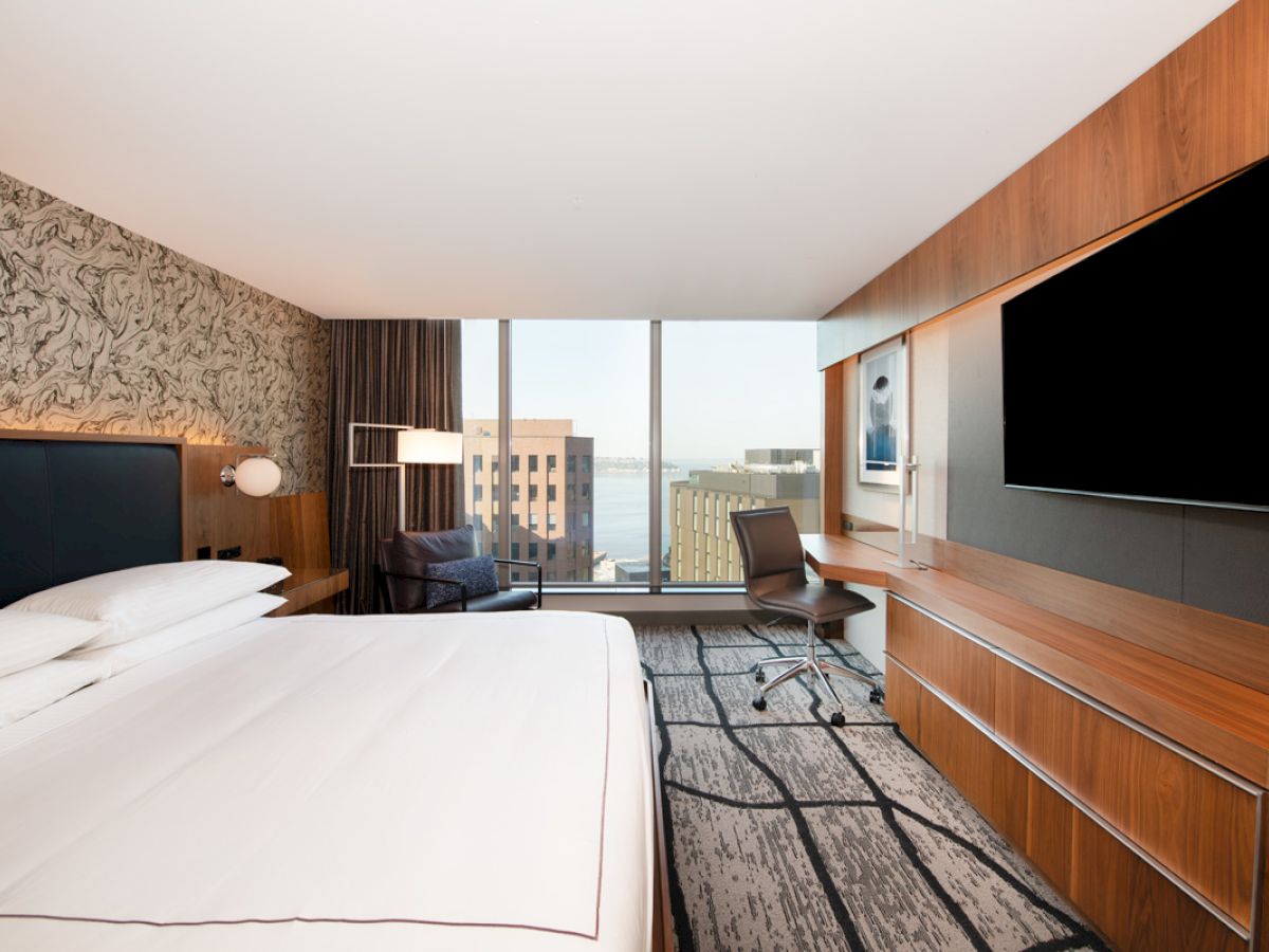 A modern hotel room with a large bed, a flat-screen TV, a desk, and a large window offering a cityscape view.