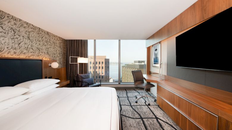 A modern hotel room with a large bed, a flat-screen TV, a desk, and a large window offering a cityscape view.