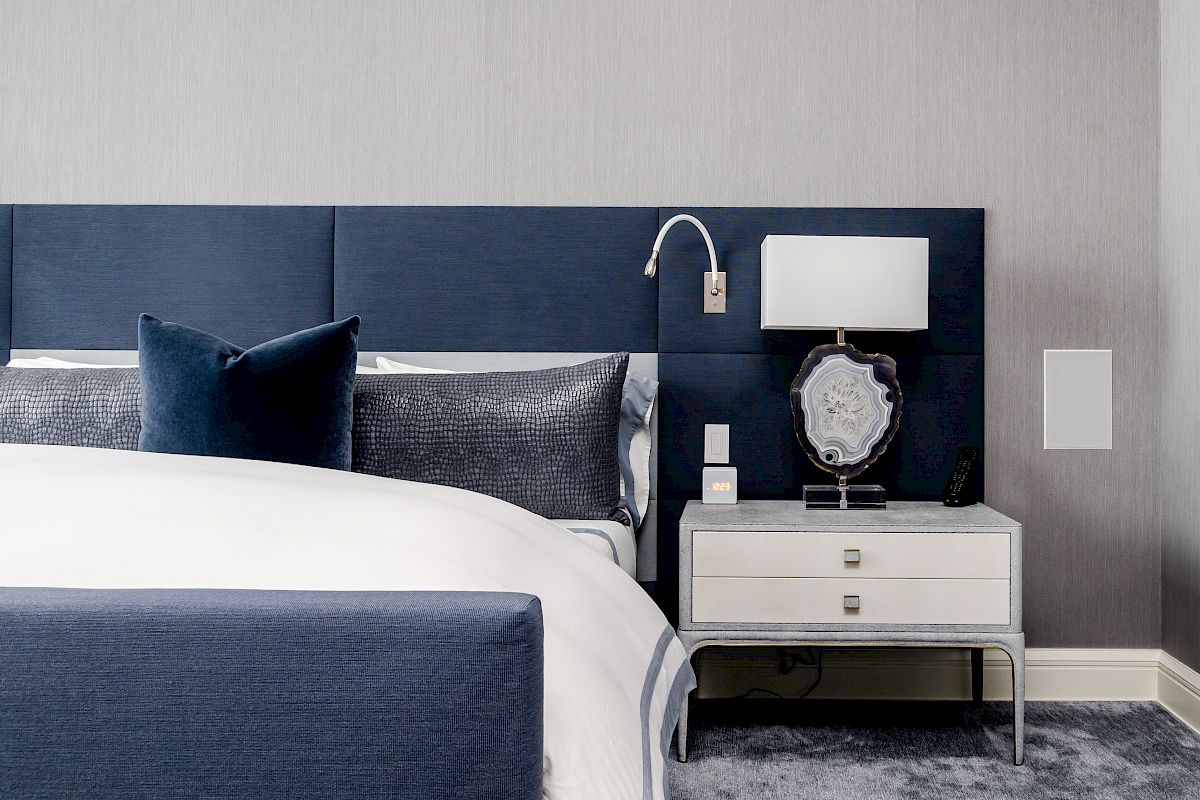 A modern bedroom with a blue and white color scheme, featuring a bed, nightstand with lamp, and wall-mounted reading light, always ending the sentence.