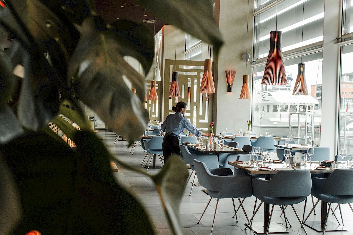 A person is setting up tables in a modern, stylish restaurant with large windows, plants, and pendant lighting.