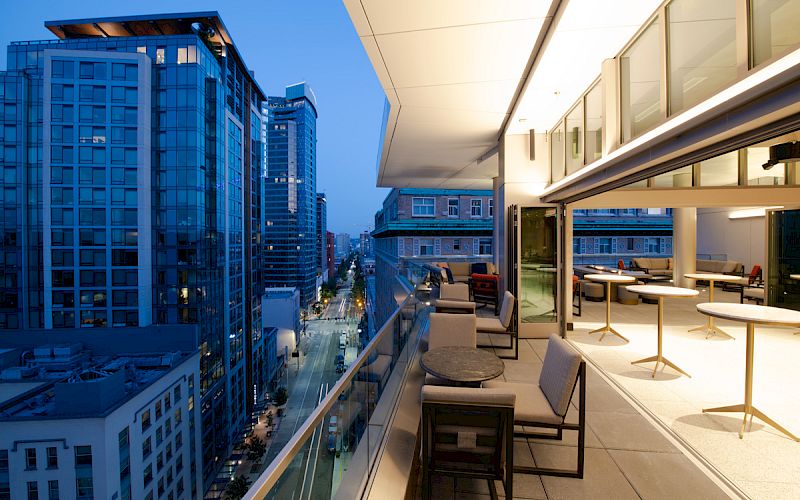 The image shows a modern cityscape, with tall buildings and a rooftop terrace featuring tables, chairs, and open seating area during evening.