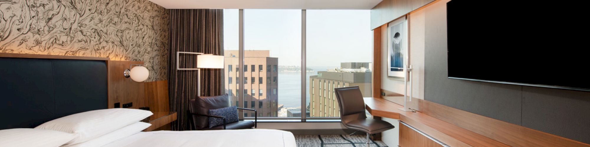 A modern hotel room with a large bed, a flat-screen TV, a desk, and a chair, featuring floor-to-ceiling windows with a city view in the background.