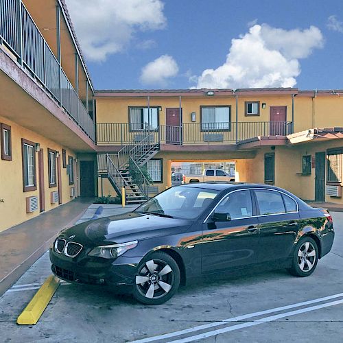 A black car is parked in front of a two-story motel with yellow walls, a vending machine, and a staircase leading up.