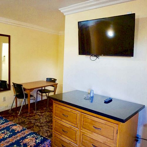 The image shows a hotel room with a bed, a wall-mounted TV, a mirror, a dresser with a remote and water bottle, and a small table with chairs.